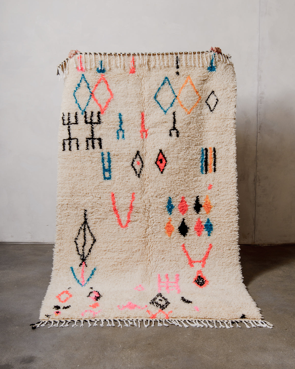 Berberlin Azilal rug made of fluffy cream wool and colourful design, held up in front of a white wall and cement floor.